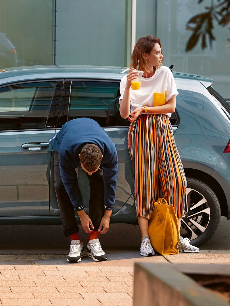 Ein Mann bindet sich seine Schuhe neben seinem Volkswagen und eine Frau trinkt Saft.