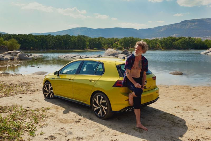 Man at the rear of the VW Golf, in the background a lake and mountains