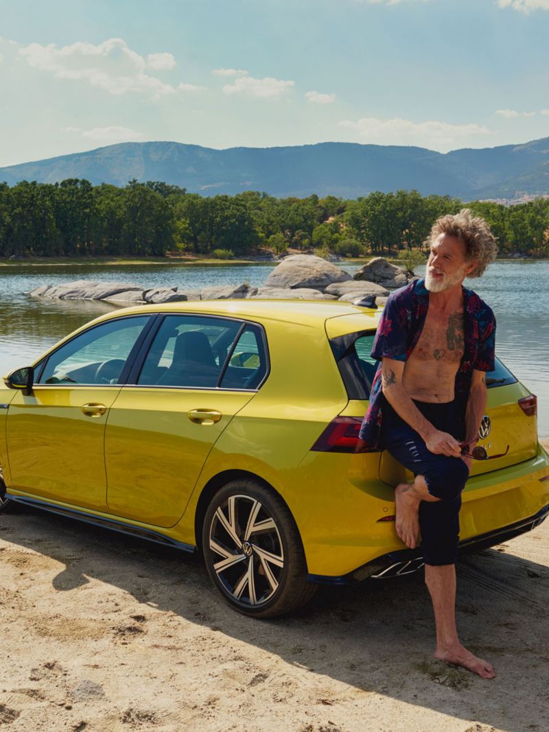 Vyras atsirėmęs į „VW Golf“ galą ežero ir kalnų kraštovaizdžio fone
