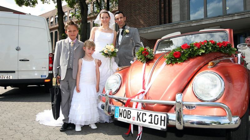 Hochzeitspaar mit zwei Kindern steht hinter einem festlich geschmückten VW Käfer (im Anschnitt), im Hintergrund steht ein Volkswagen Transporter