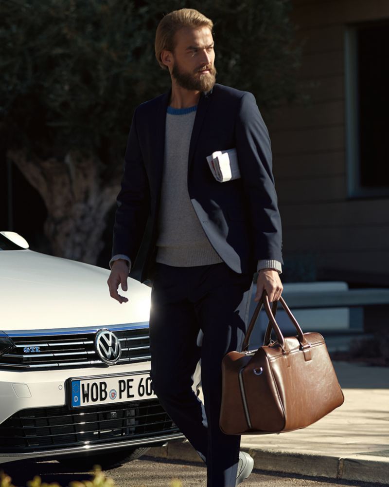 Geschäftsmann geht mit einer Tasche in der Hand vor einem weißen Volkswagen Passat GTE entlang.