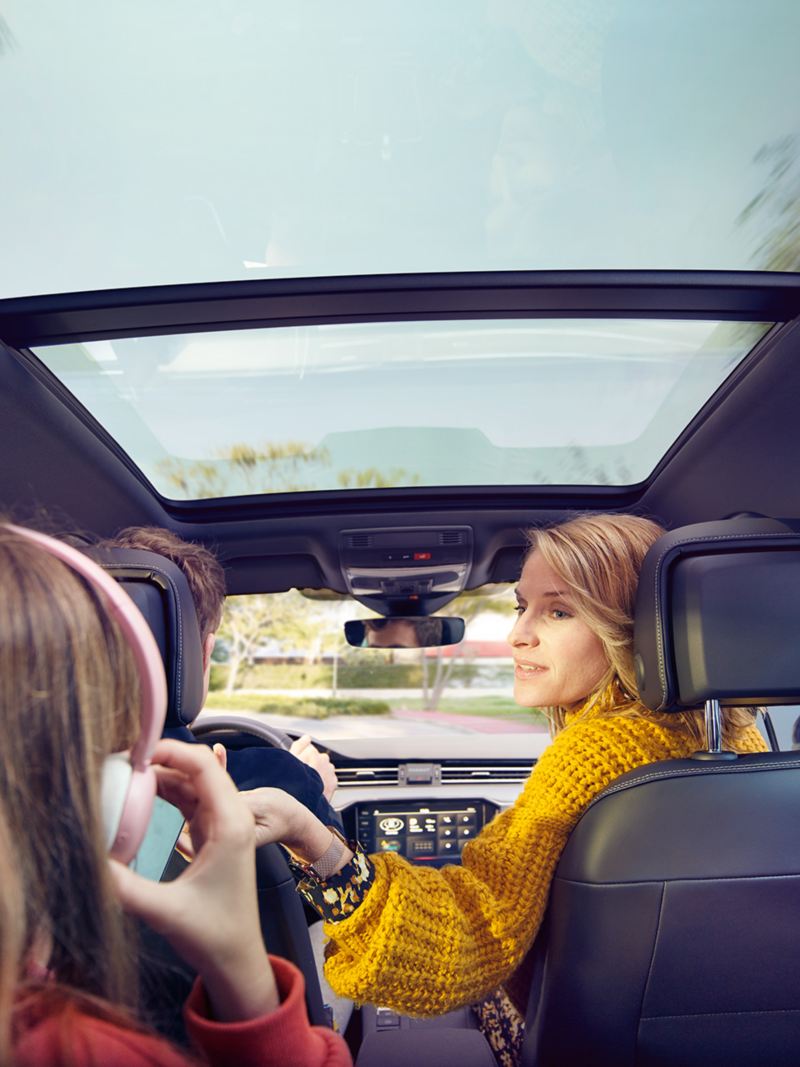 Vehicle interior of the VW Passat Estate with optional panoramic sunroof. Woman in the front passenger seat turns round to a child on the rear bench seat. 