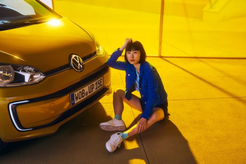 Una ragazza siede per terra di fronte a Volkswagen Nuova e-up!, vista frontalmente con dettaglio sul cofano.