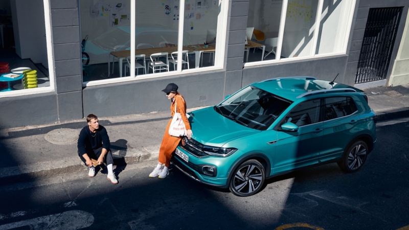 Volkswagen T-Cross bleu turquoise garé dans une rue avec une femme assise sur le capot et un homme assis devant le véhicule