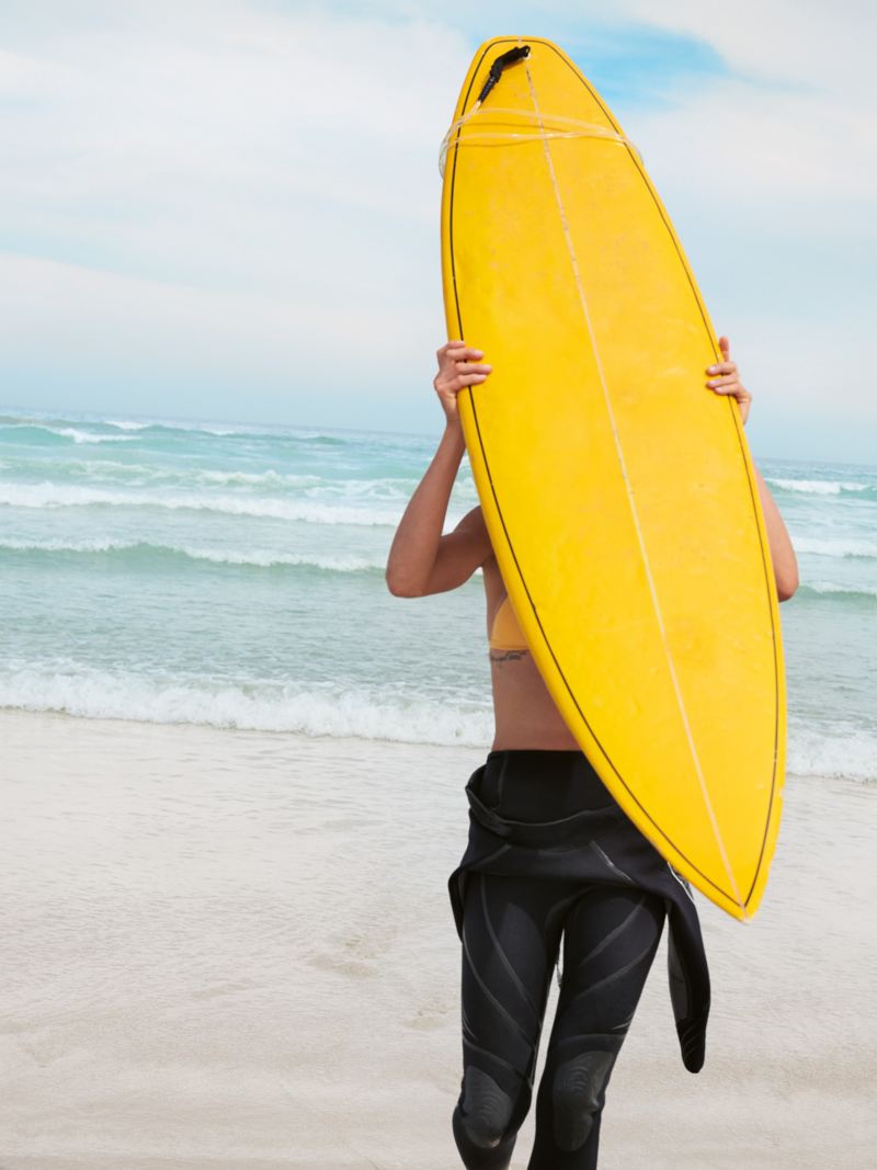 En hvid sandstrand med hav, en kvinde går hen mod den og bærer et surfbræt på ryggen.