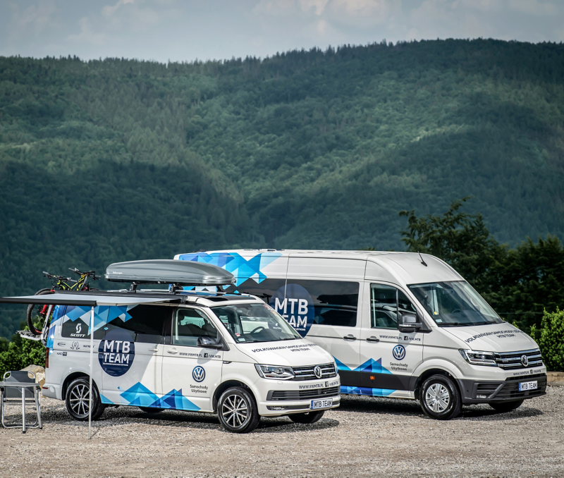 Volkswagen Crafter oraz Volkswagen California oklejone w logo MTB Team stoją na tle górzystego lasu