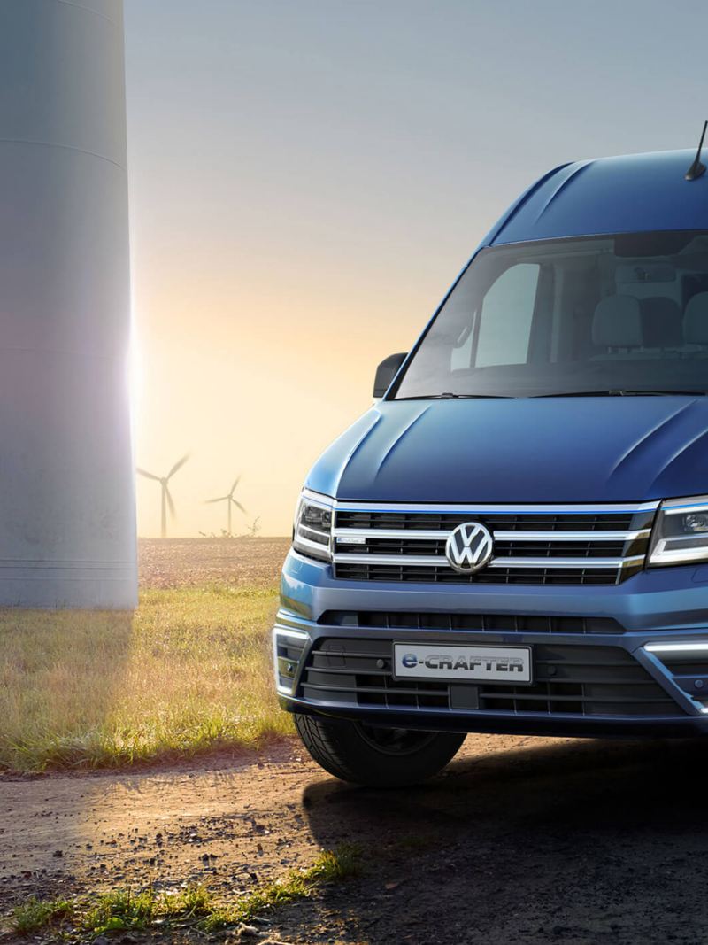 Blue VW e-Crafter front view with a grassy field and wind turbines in the background