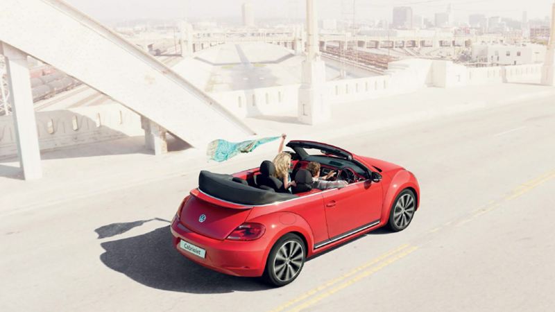 A couple driving a red Volkswagen Beetle Cabriolet, the roof down next to an old city.