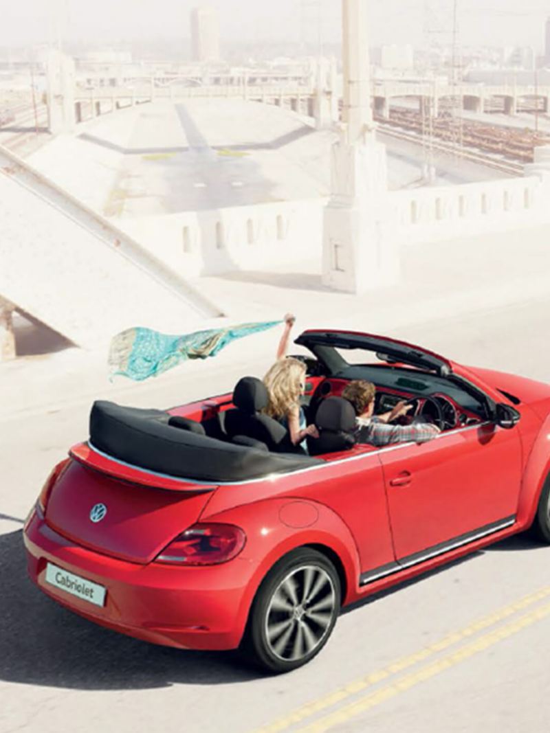 A couple driving a red Volkswagen Beetle Cabriolet, the roof down next to an old city.