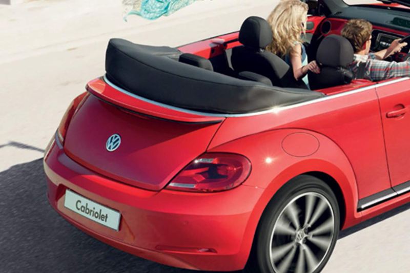 Rear shot of a red Volkswagen Beetle Cabriolet, a couple driving next to the beach with the roof down.