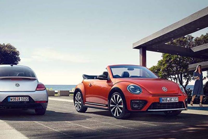 A orange Volkswagen Beetle Cabriolet, parked next to a beach on a promenade.