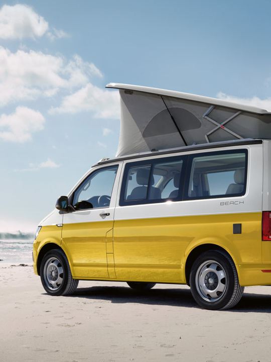 En gul Volkswagen California holder på stranden i solskinsvejr. To surfere løber hen imod den.