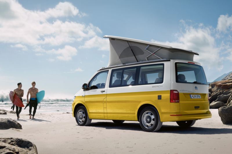 En gul Volkswagen California holder på stranden i solskinsvejr. To surfere løber hen imod den.
