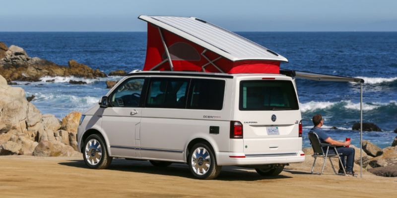En Volkswagen California med højt rødt tag holder på stranden. Markisen er kørt ud. Føreren sidder under markisen og kigger på havet.