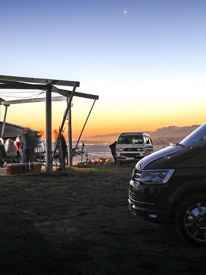 Mehrere unterschiedliche Volkswagen Nutzfahrzeuge California-Modelle stehen bei Sonnenuntergang an einer Küste. Im Hintergrund kann man das Meer erahnen.