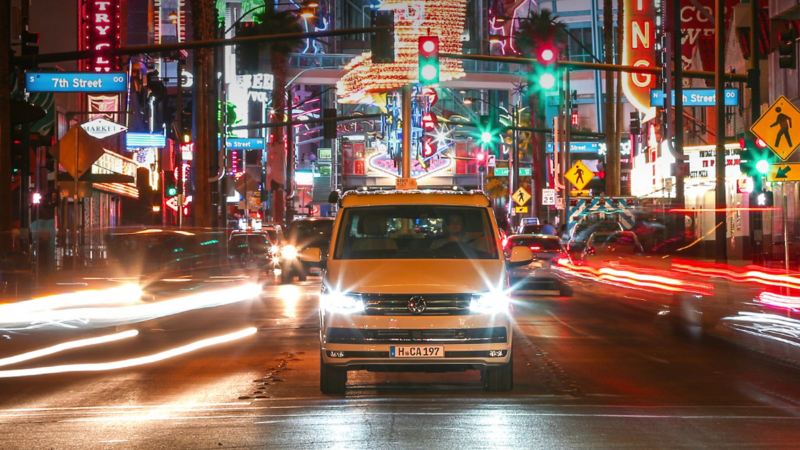 Der Volkswagen Nutzfahrzeuge California fährt durch eine kalifornische Innenstadt. Um ihn herum funkeln bunte Leuchtreklamen.