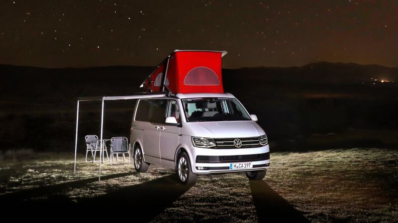 Der Volkswagen Nutzfahrzeuge California steht bei Nacht auf einer Wiese. Die Markise ist ausgefahren. Darunter stehen Campingmöbel.