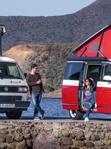 To Volkswagen California-modeller med højt tag holder på en mole ved havet.