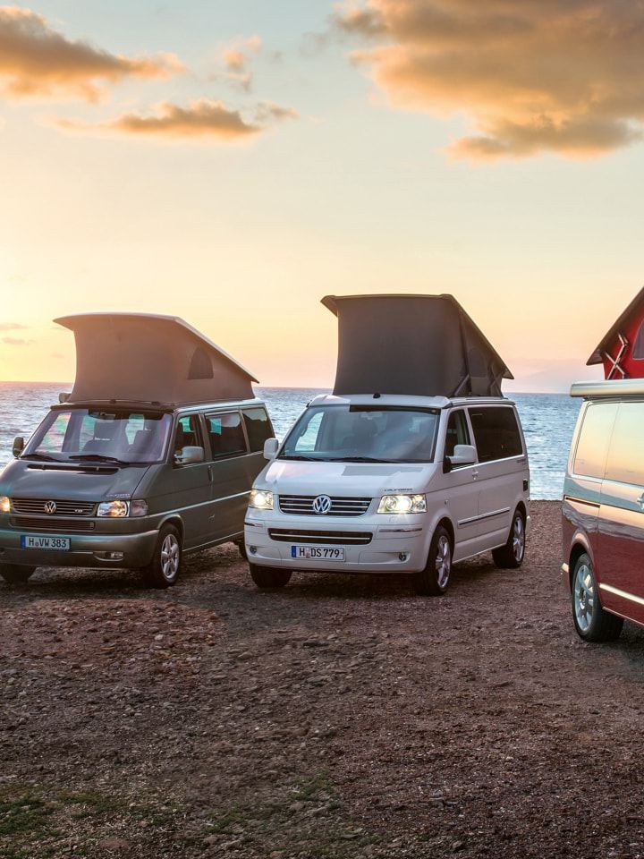 Fire Volkswagen California-modeller fra forskellige generationer holder på stranden ved solnedgang.