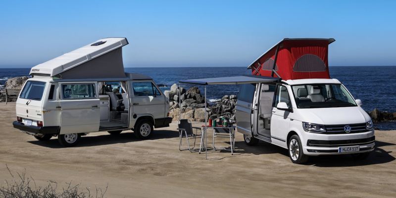 Zwei Volkswagen Nutzfahrzeuge California mit Hochdächern stehen an einem felsigen Strand. Beim rechten Fahrzeug ist die Markise ausgefahren. Darunter stehen Campingmöbel.