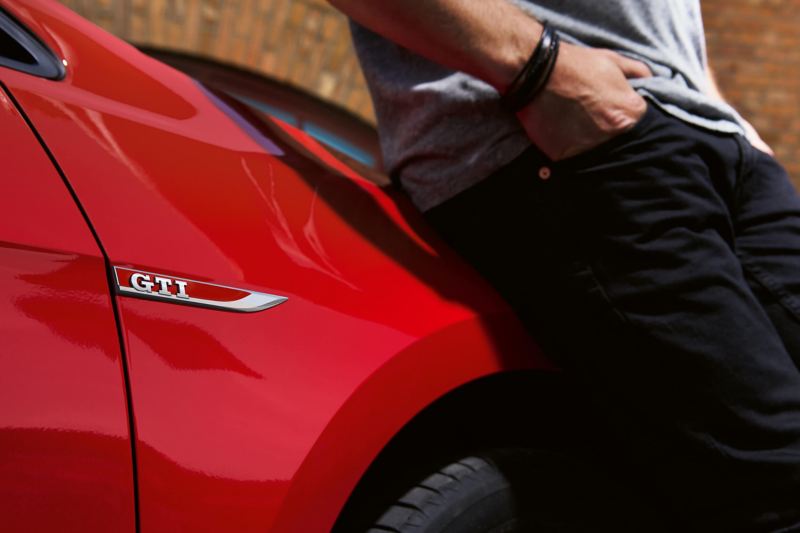 Man leaning on the front side of a red Volkswagen Golf GTI, logo in shot above the front wheel.