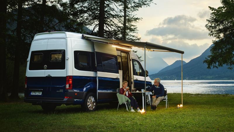 Le Grand California le soir, devant un lac de montagne. Un couple d'un certain âge est assis sous l'auvent, à la table de camping, et boit un thé, sous un éclairage agréable.