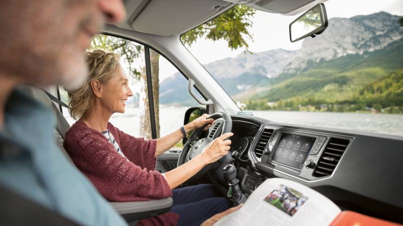 Sguardo sulla plancia di Volkswagen Grand California dal lato passeggero. Una donna siede al volante, il suo compagno è sul sedile lato passeggero. Sullo sfondo si vede un panorama montano con un lago. 