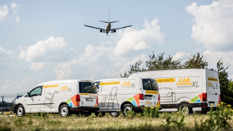 Caddy, Transporter i Crafter w okleinie o carsharingu stoją na polanie, nad nimi leci samolot