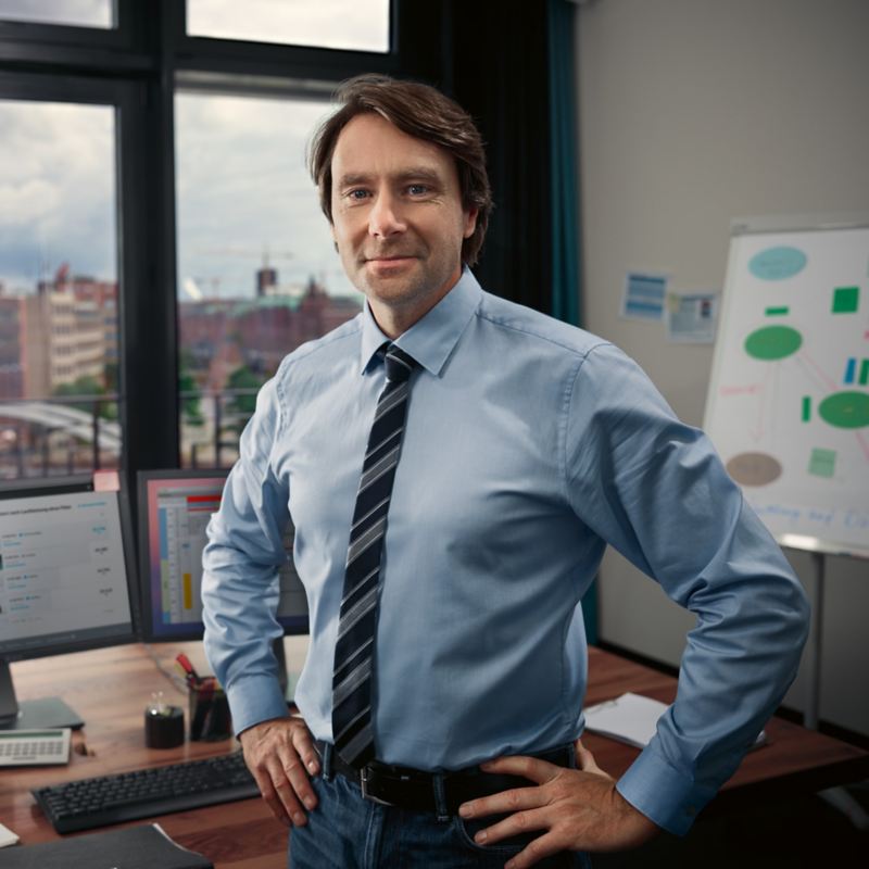 Ein Mann im Hemd steht vor einem Schreibtisch mit den Händen in die Hüfte gestützt