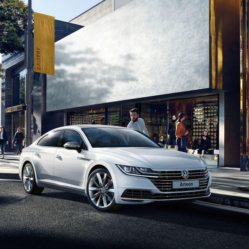 A white Volkswagen Arteon parked on a city street outside of shops, the owner getting in.