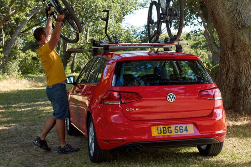 A man hitching his bike to the roof rake of a Volkswagen Golf, surrounded by woodland