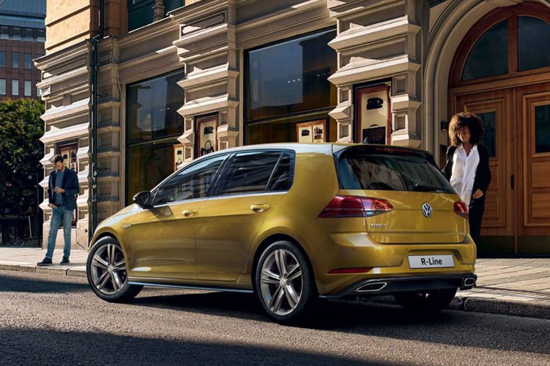 A gold Volkswagen Golf R-Line, parked outside an old city building, with a lady approaching. 