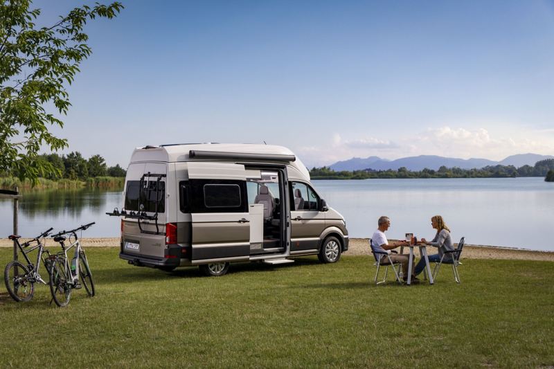 Volkswagen Grand California i para przy stoliku nad jeziorem