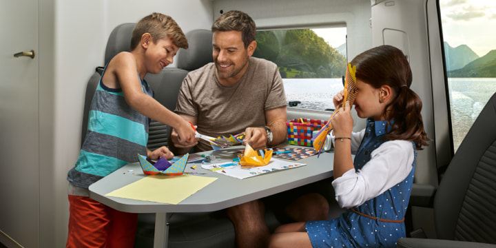 Ein Vater und seine Kinder sitzen im Wohnbereich des Grand California am Tisch und spielen. Im Hintergrund ist die Mutter zu sehen. Sie liegt im Bett und liest ein Buch.