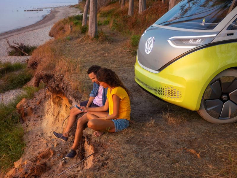 Volkswagen ID.Buzz kuvattuna rantatörmällä, auton keula näkyy kuvassa ja auton edessä on mies ja nainen selaamassa älypuhelinta ilta-auringon valaisemana