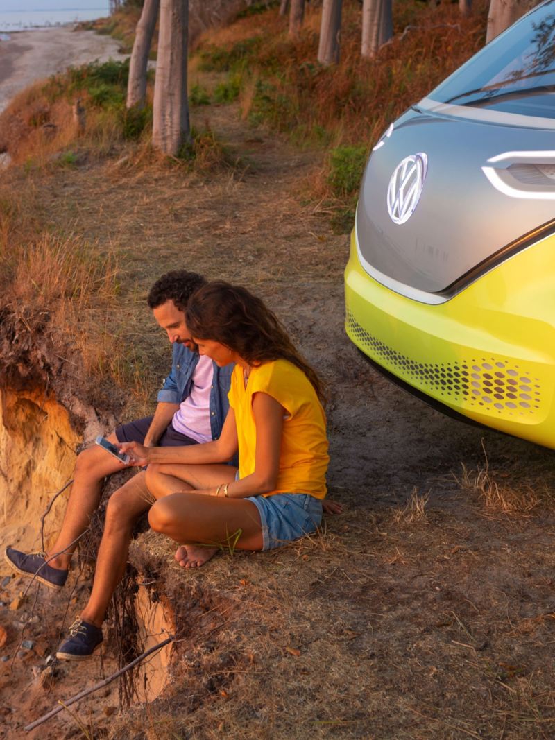 Volkswagen ID.Buzz kuvattuna rantatörmällä, auton keula näkyy kuvassa ja auton edessä on mies ja nainen selaamassa älypuhelinta ilta-auringon valaisemana