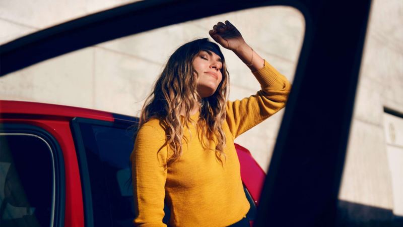Kvinna som står i solen vid en Volkswagen och har ansiktet vänt mot solen och håller handen framför ansiktet för att inte bli bländad