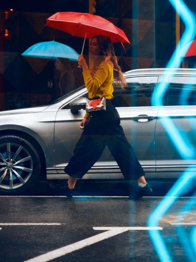 Kvinna med rött paraply går på en väg. Volkswagen bil syns i bild.