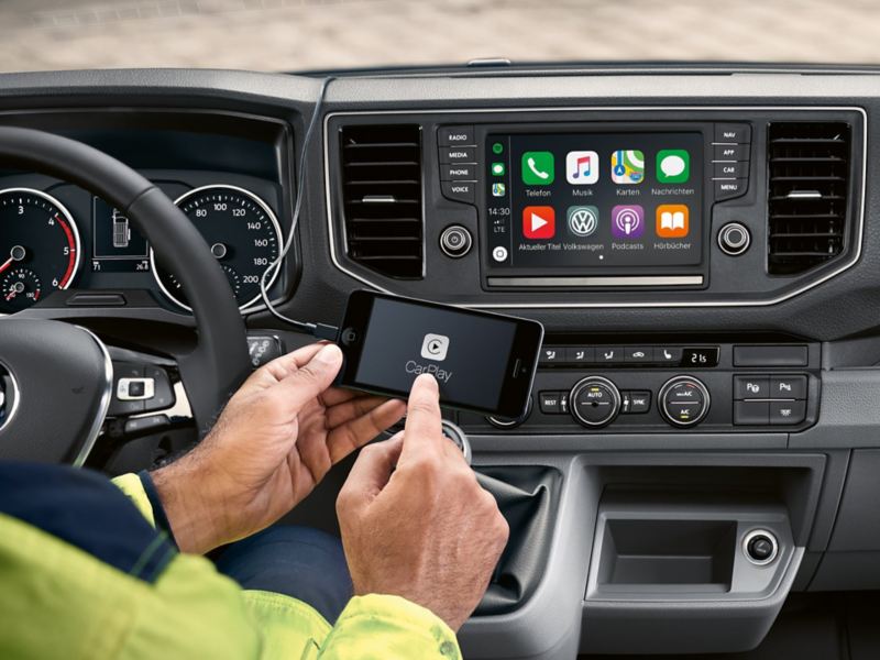 Intérieur de la cabine du Crafter à plateau à l’arrêt. En arrière-plan, le service en ligne Car-Net apparaît sur l’écran multifonctions. Au premier plan, un homme utilise son smartphone.