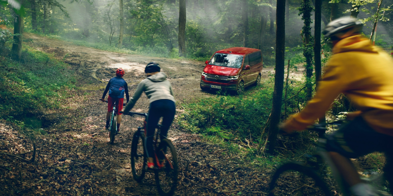 Drei Personen fahren im Wald einen Weg runter. Im Hintergrund ein roter Multivan 6.1.