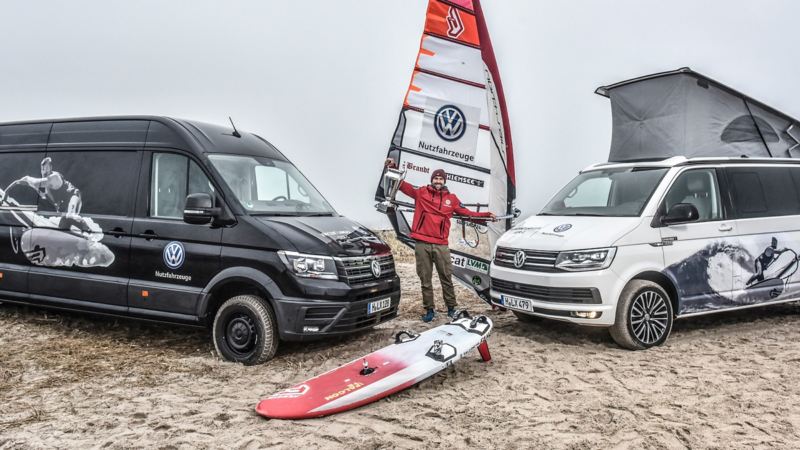 Vincent Langer steht mit seinem Windsurf-Board zwischen zwei Multivans am Strand.