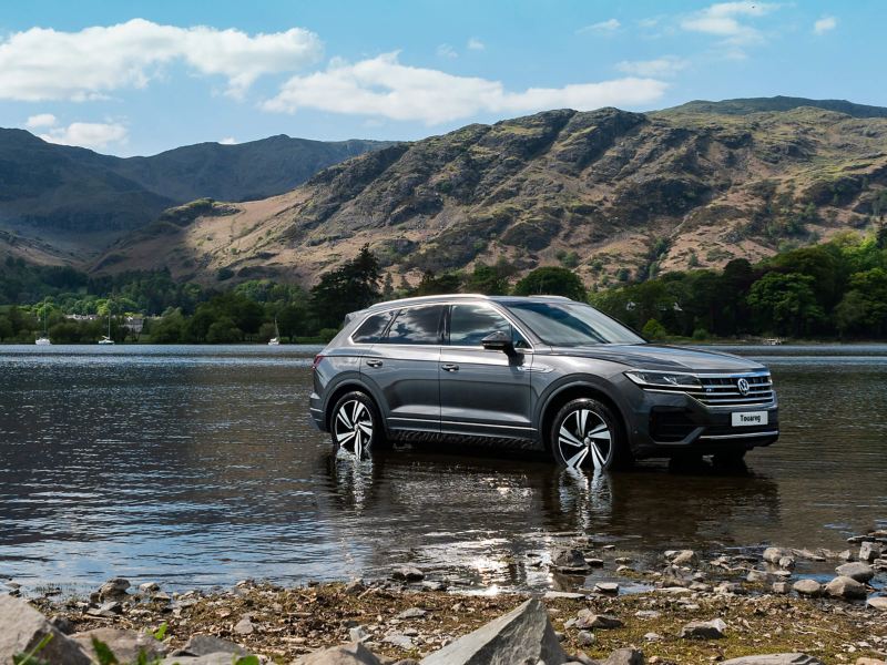 A slate silver Volkswagen Touareg wading through a shallow lake, surrounded by mountains.