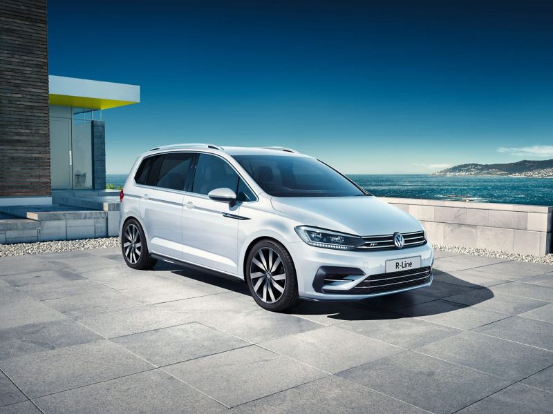 A white Volkswagen Touran outside a building with a bay and white cliffs in the background.