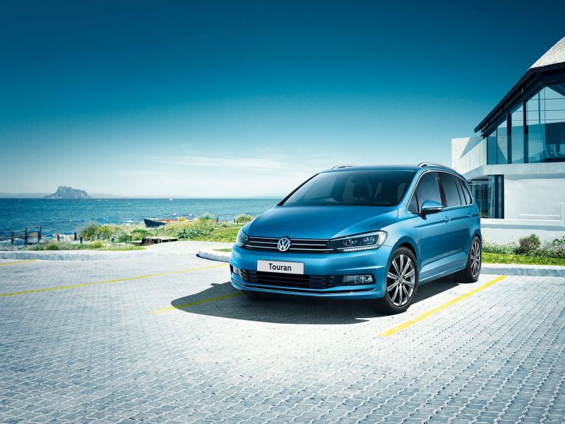 A blue Volkswagen Touran outside a beachfront house, the sea in the background.
