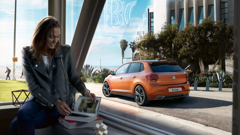 A lady reading a book in a cafe with a view through the window of the Polo R-Line parked near the coast facing out to sea