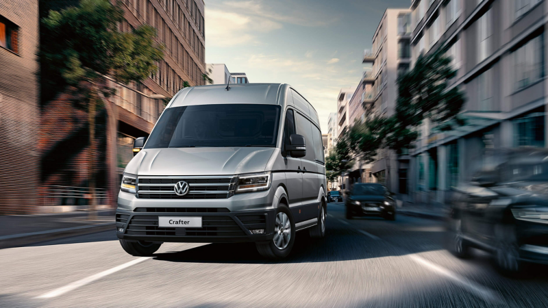 A silver Volkswagen Crafter van driving through a modern city street, surrounded by blurred buildings, trees, and other vehicles, creating a sense of motion. The front grille displays the Volkswagen logo, and the van’s headlights are on. The urban setting features contemporary architecture with large windows and greenery.