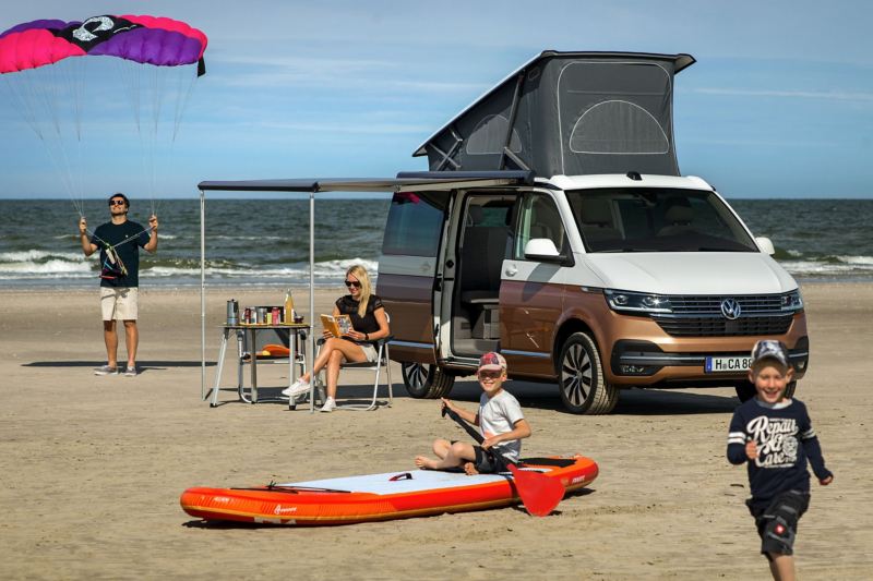 Rodzina bawi się na plaży, obok stoi Nowa California 6.1 z rozłożonym dachem sypialnym i markizą