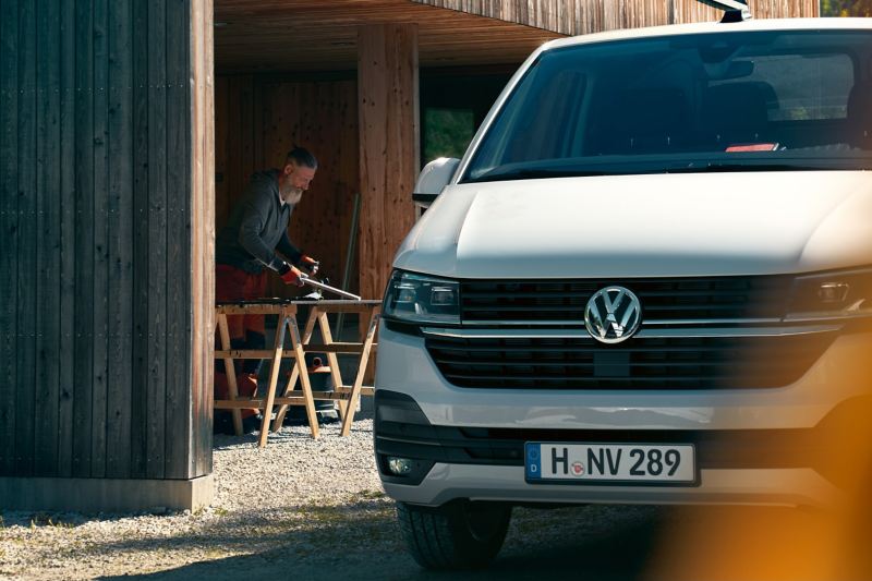 Ein Volkswagen Transporter 6.1 Kastenwagen vor einer Holzwerkstatt. Im Hintergrund arbeitet ein Schreiner.
