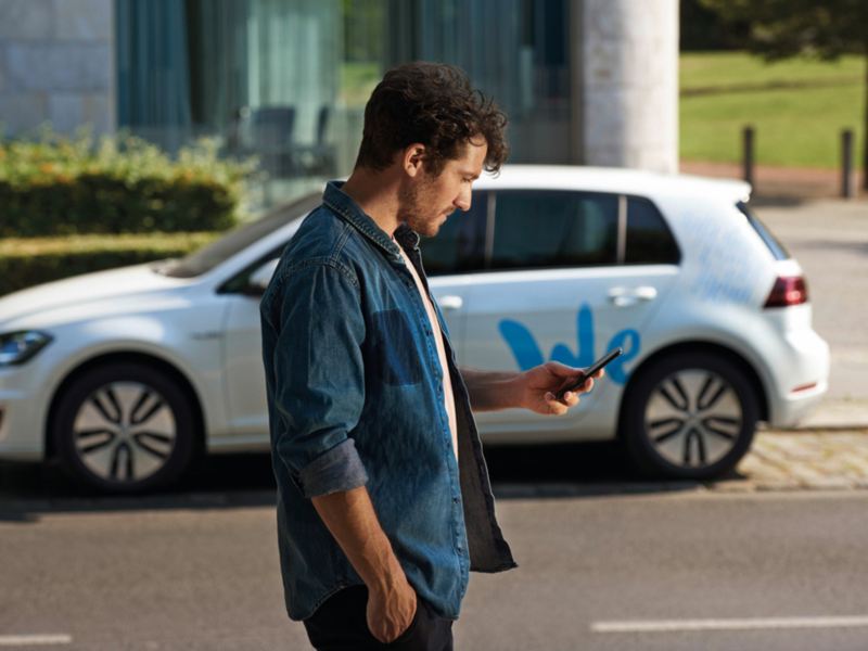 Hombre de pie en una calle mirando al móvil y de fondo un coche aparcado con la palabra WE estampada