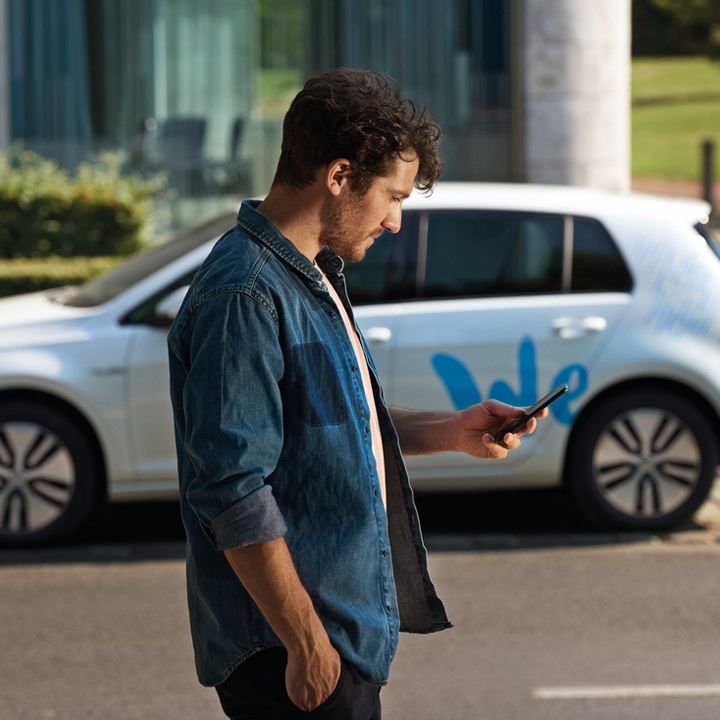 Hombre joven consultando un móvil delante de un Golf blanco aparcado con el logo de Volkswagen We